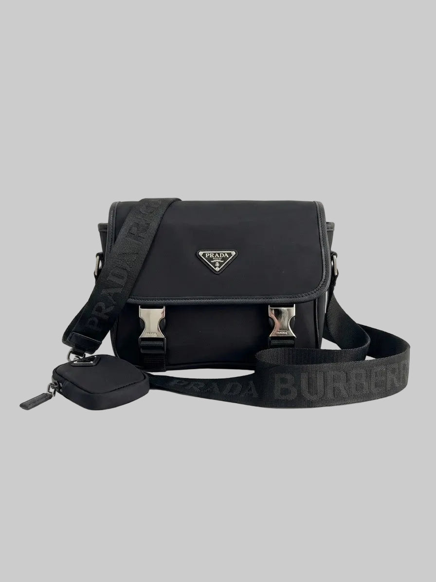 A black Prada shoulder bag with a matching zippered pouch and a wide black Burberry strap, displayed on a clean white reflective surface.